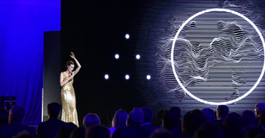 Na zdjęciu śpiewająca kobieta w złotej sukni, obok, na ekranie, widać wielkie koło