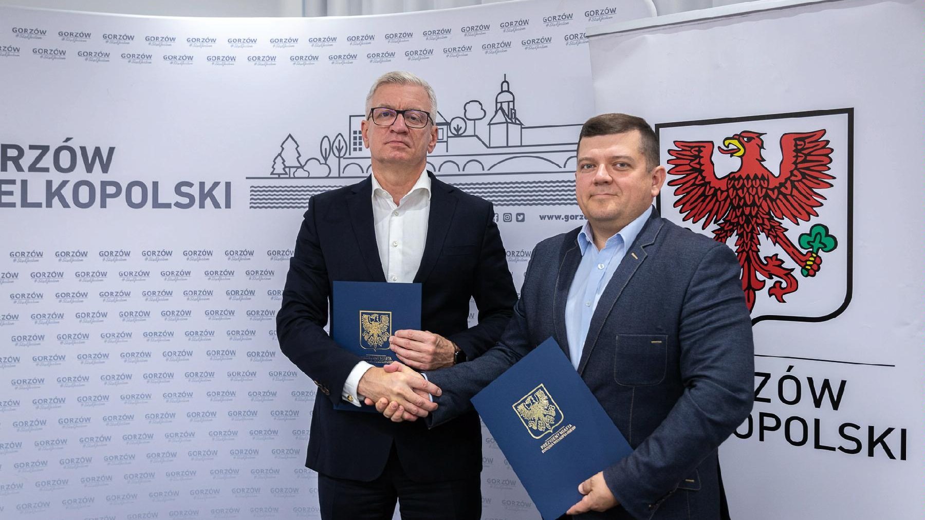 Prezydenci Jacek Jaśkowiak i Jacek Wójcicki stoją obok siebie, podając sobie dłonie. W rękach mają teczki z podpisanym porozumieniem. W tle - ścianka z herbem Gorzowa Wielkopolskiego - grafika artykułu