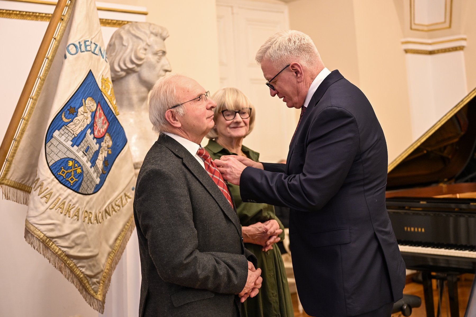 Zdjęcie przedstawia prezydenta przypinającego medal kobiecie. Obok stoi mężczyzna. Widać też flagę z herbem Poznania.