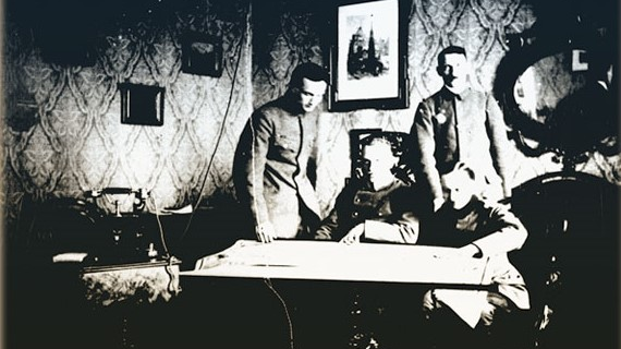 Trzech mężczyzn w mundurach pochyla się nad stołem w mieszkaniu.