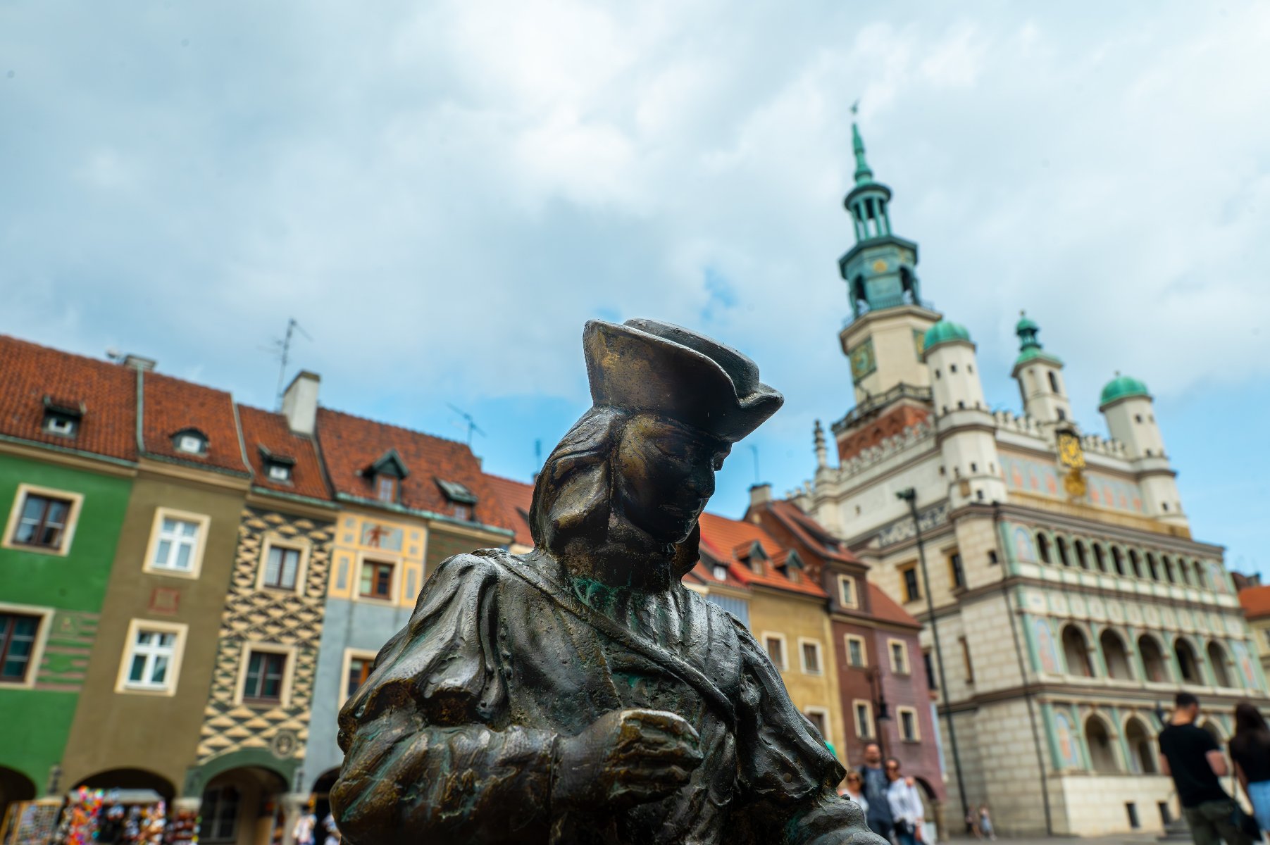 Brązowa rzeźba przedstawia siedzącego starszego mężczyznę w kapeluszu. W tle kolorowe kamienice Starego Rynku i zielona wieża Ratusza. Niebo jest częściowo zachmurzone.