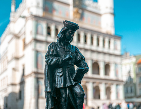 Czarna rzeźba w kształcie postaci w historycznym stroju, trzymającej zwój i pióro, na pierwszym planie. W tle, zamazany, biały renesansowy Ratusz w Poznaniu.