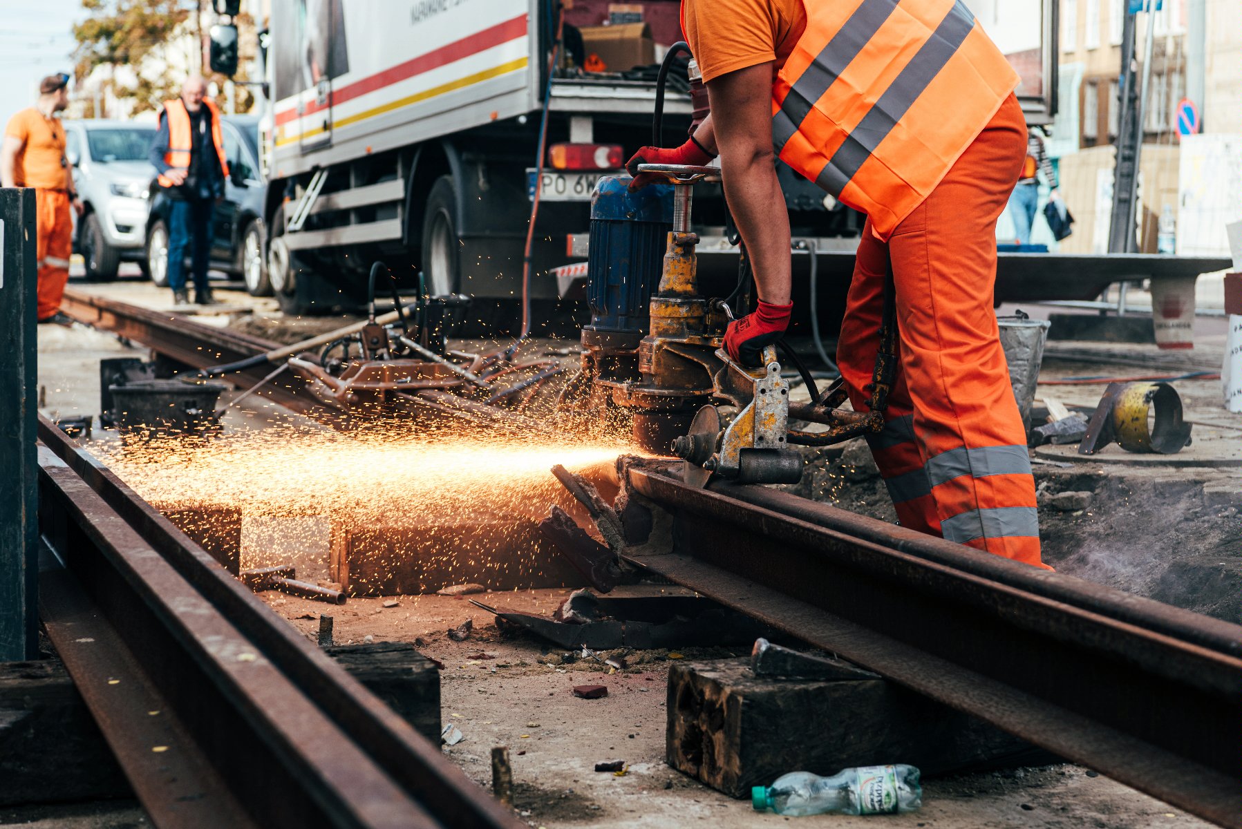 Naprawy odbedą się w nocy. Na zdjęciu widać robotknika przycinającego metalowe elementy. Nachyla się nad nimi. Widać jak lecą iskry z przycinnanego elementu. W tle plac budowy z ciężarówkami.