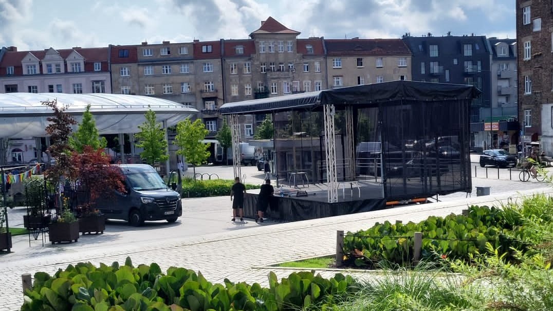 Po prawej stronie widok na pracowników, który montują scenę koncertową. Po lewej stoi bus monterów. W tle budynki kamienic przy Łazarskim Rynku i widoczne okrągłe zadarzenie nad powierzchnią handlową rynku.