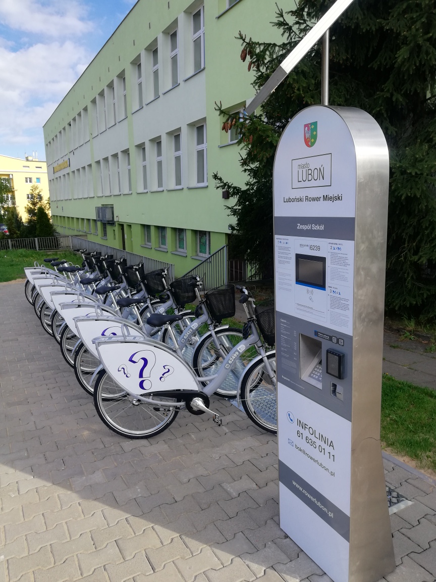 8 rowerów miejskich ustawionych przed zielonym budynkiem. Po prawej automat stacji rowerowej. - grafika artykułu