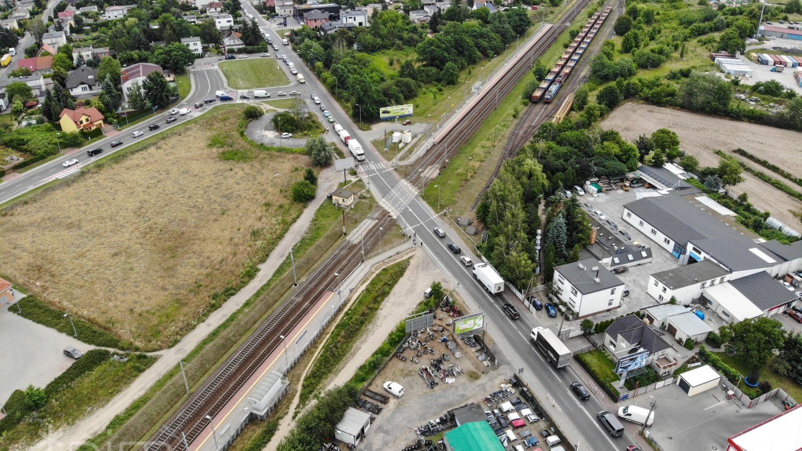 Ul. Grunwaldzka i tory kolejowe. Widok z góry. Długa kolejka samochodów z obu stron zamknietego przejazdu kolejowego. W lewym górnym i prawym dolnym rogu budynki.