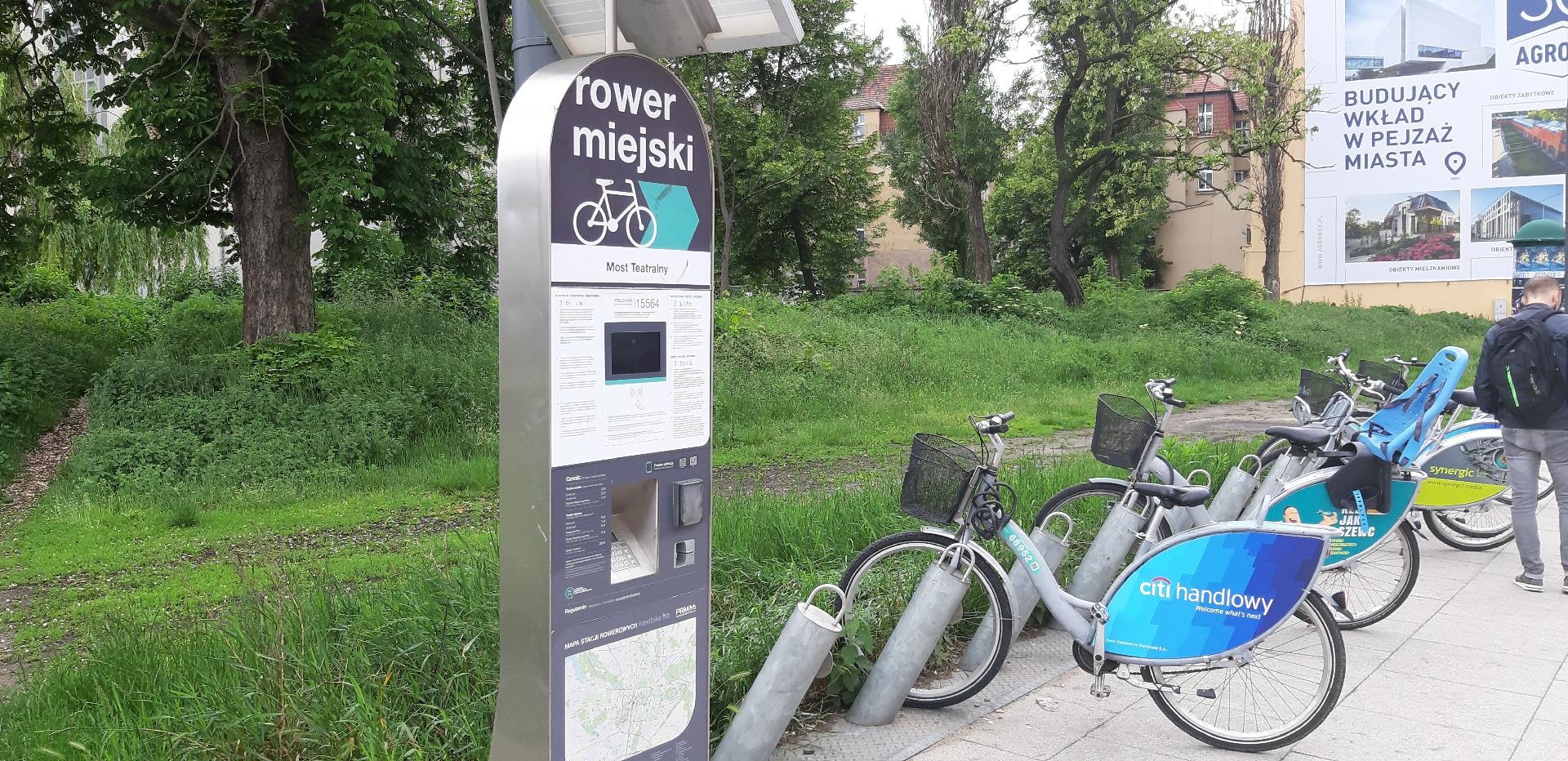 Stacja Poznańskiego Roweru Miejskiego, zaparkowany rower. Wyżej drzewa i teren zoielony. - grafika artykułu