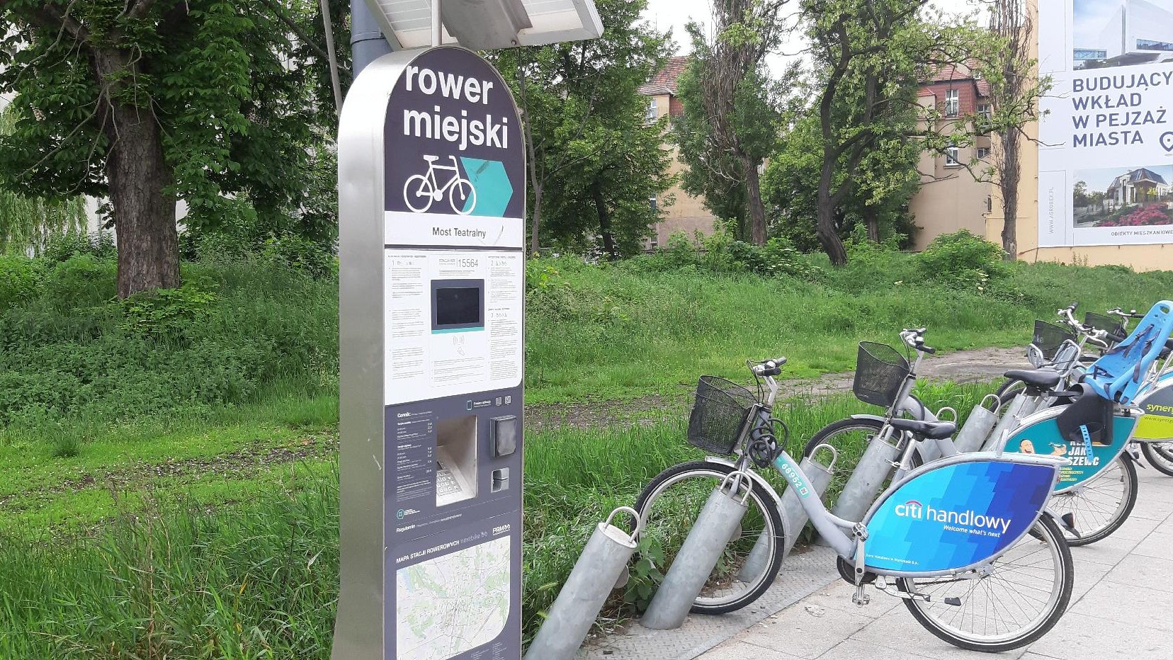 Stacja Poznańskiego Roweru Miejskiego, zaparkowany rower. Wyżej drzewa i teren zoielony. - grafika artykułu
