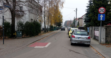 Widok na ulicę z kontraruchem rowerowym. Po lewej domy. Po prawej zaparkowane samochody.