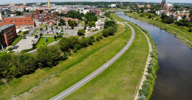Wartostrada. Widać na nim drogę rowerową prowadzącą przez tereny zieleni. W sąsiedztwie znajdują się się zabudowania i rzeka.