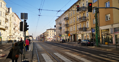 Sygnalizacja świetlna przy ul. Kanałowej. Po lewej piesi na przystanku tramwajowym. Po prawej zaparkowany samochód i rowerzysta.