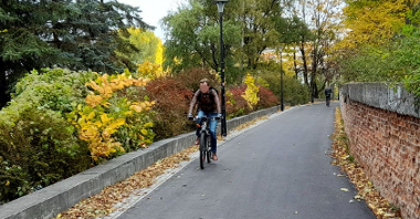 Zdjęcie przedstawia asfaltową ścieżkę pieszo rowerową. Wokół dużo drzew, bliżej obiektywu mężczyzna na rowerze, dalej w tle pieszy.