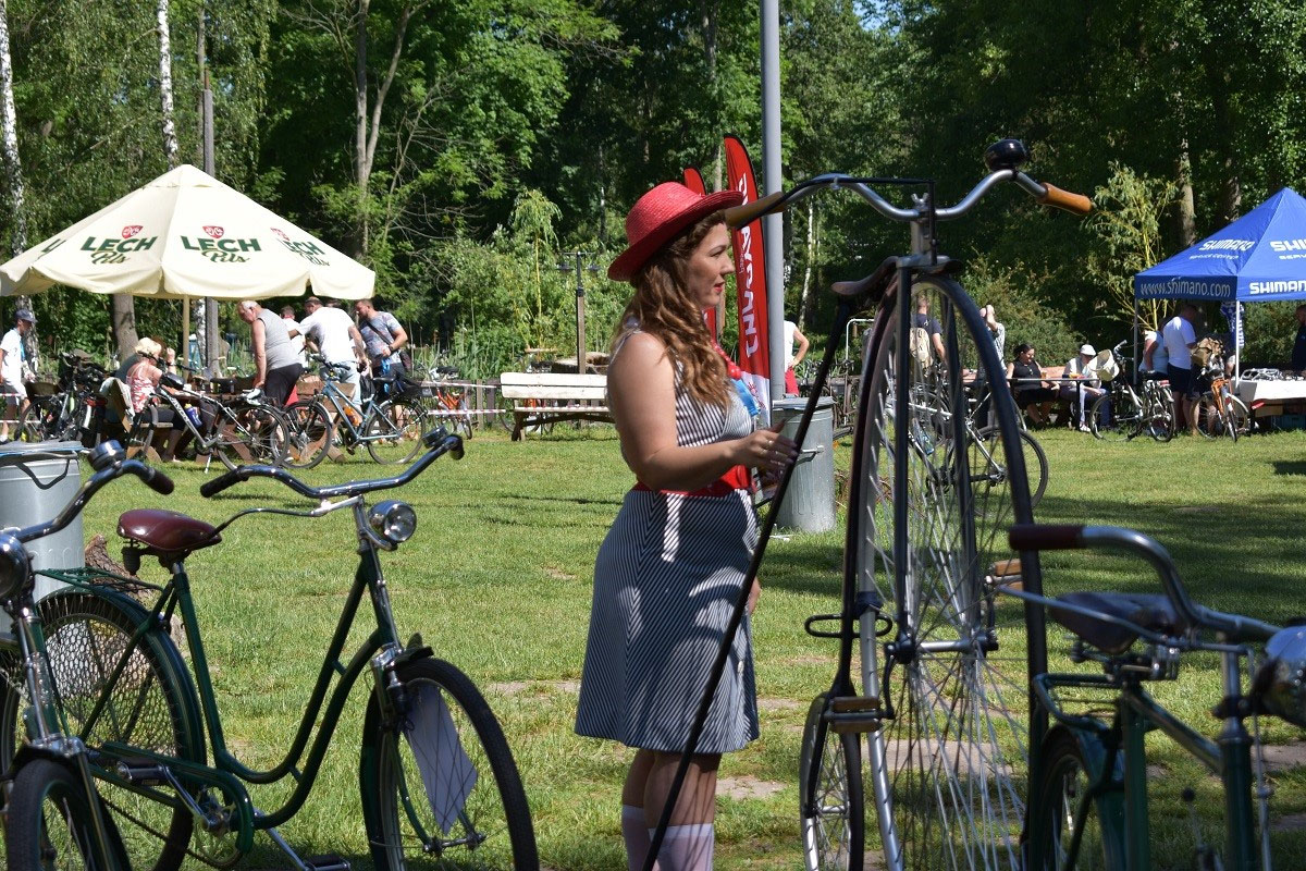Bicykl a obok niego stary rower bez ramy, tzw. damka. Obok bicykla kobieta w kapeluszu. W głębi parasole, stoły, ludzie i rowery. - grafika artykułu