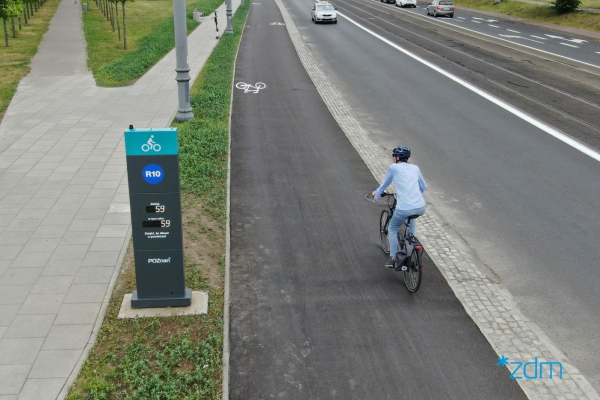 Widok z góry na drogę rowerową na ul. Kórnickiej. Rowerzystka ubrana na niebiesko w kasku jedzie po drodze rowerem. Po lewej stronie licznik rowerowy. Po prawej droga dla samochodów i tramwaji. - grafika artykułu