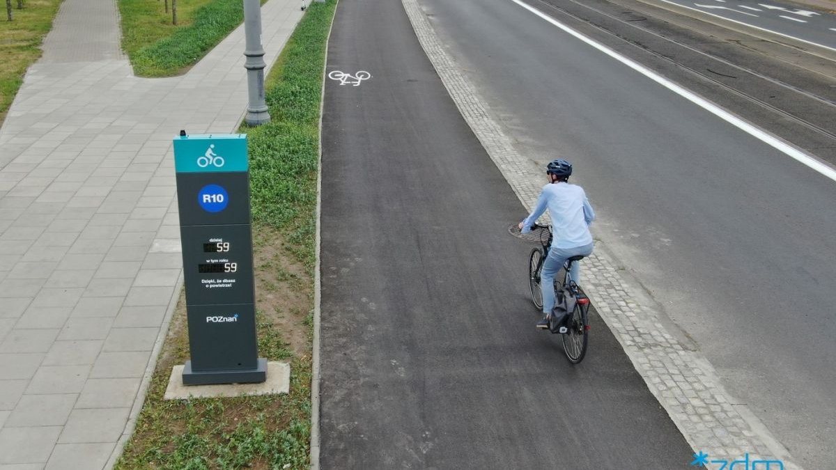 Widok z góry na drogę rowerową na ul. Kórnickiej. Rowerzystka ubrana na niebiesko w kasku jedzie po drodze rowerem. Po lewej stronie licznik rowerowy. Po prawej droga dla samochodów i tramwaji. - grafika artykułu