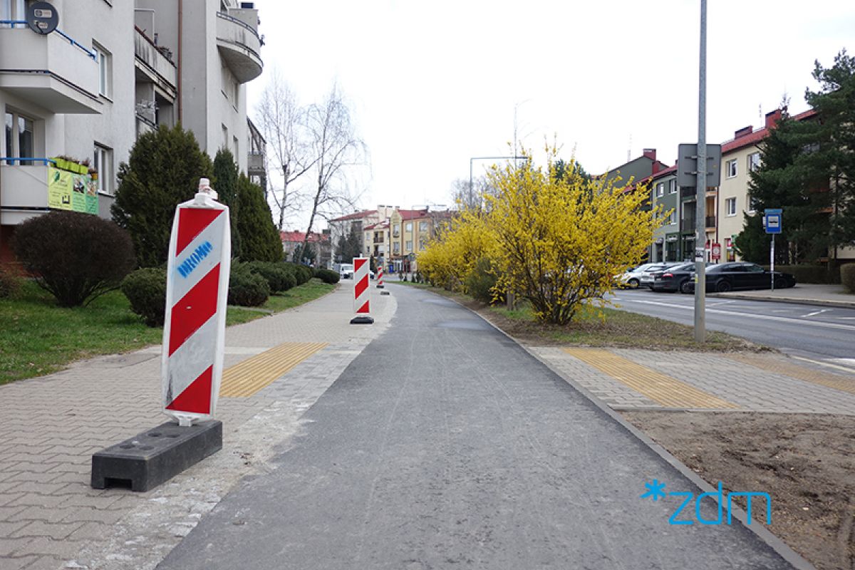 Na zdjęciu widać nowo wybudowaną drogę rowerową w terenie zabudowanym. Wzdłuż niej po jednej stronie ciągną się żółte krzewy, a po drugiej pachołki ostrzegawcze. - grafika artykułu
