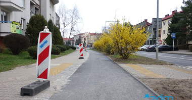 Na zdjęciu widać nowo wybudowaną drogę rowerową w terenie zabudowanym. Wzdłuż niej po jednej stronie ciągną się żółte krzewy, a po drugiej pachołki ostrzegawcze.