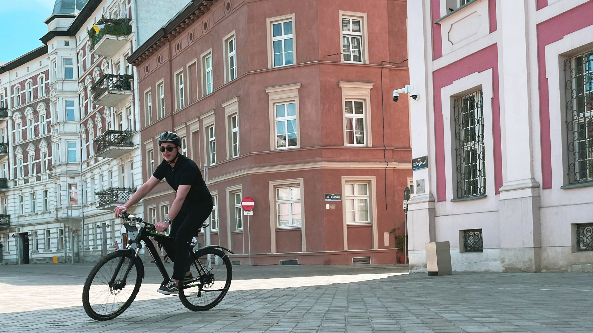 Ubrany na czarno rowerzysta w kasku jedzie przez oświetlony promieniami słońca plac Kolegiacki. W tle widać budynki, pomiędzy którymi znajduje się zjazd w uliczkę. - grafika artykułu