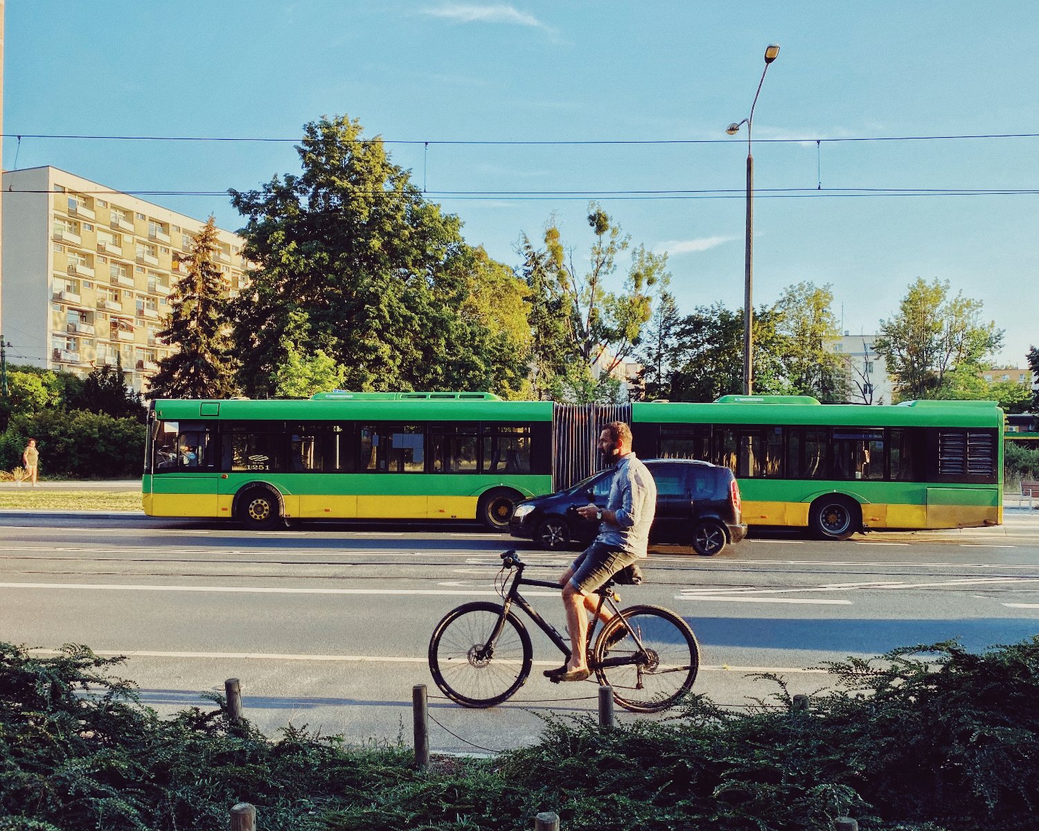 Na obrazku widać rowerzystę na pierwszym planie, na kolejnym samochód, a w oddali autobus. Świeci słońce. Zdjęcie obfituje w zieleń. - grafika artykułu