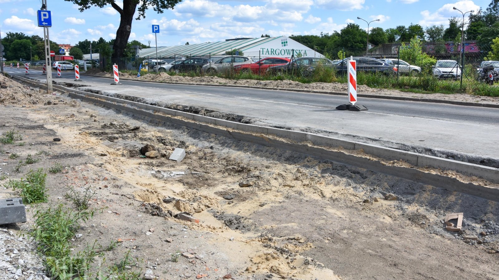 Po lewej rozkopany teren, na srodku jezdnia podzielona na dwie części szykanami biało-czerwonymi. Po prawej hala targowa i zaparkowane samochody.