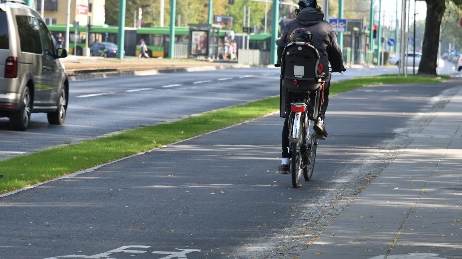 Rowerzysta z dzieckiem na krzesełku rowerowym jedzie drogą rowerową. Na jezdni obok samochód. Dalej widać tramwaj. - grafika artykułu