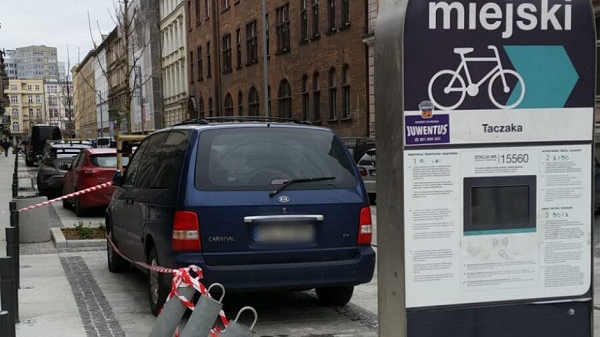 Stojaki rowerowe i zaparkowane samochody. Po prawej kamienice.