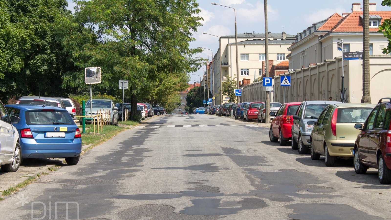 Ulica ze zniszczoną nawierzchnią, po obu jej stronach gęsto zaparkowane samochody, także na trawniku