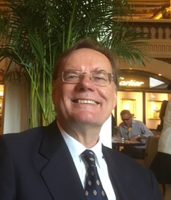 A smiling man in a dark jacket stands against a background of trees