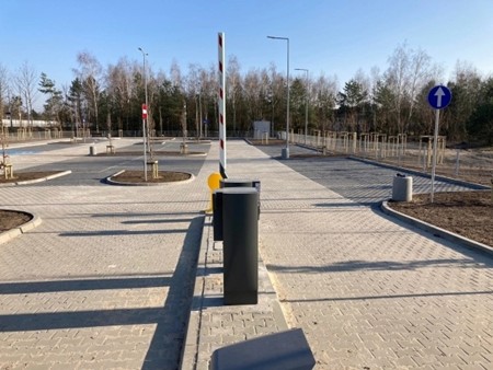 At the centre there is an entry barrier with a parking payment machine. The surface is made of paving stones, with clearly marked traffic lanes and parking spaces. On the right-hand side, a mandatory one-way traffic sign is visible. The car park is empty, with no vehicles parked.