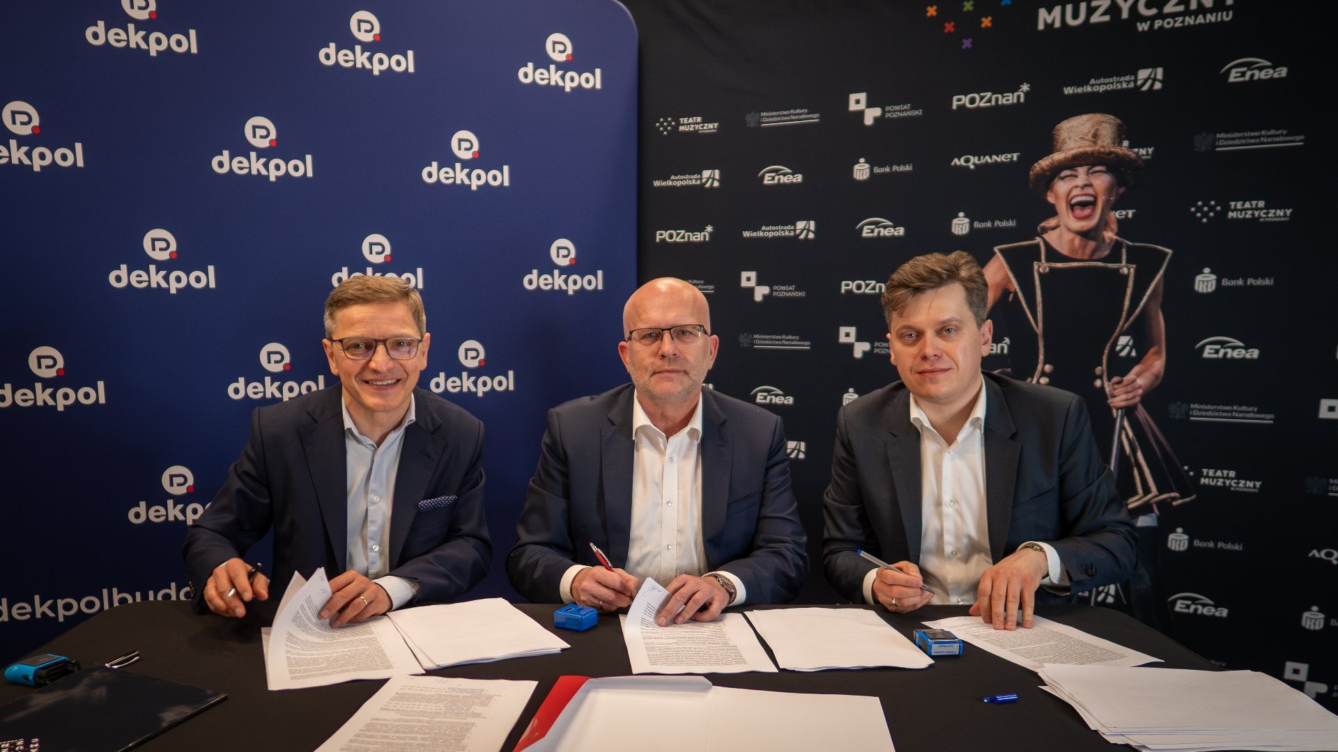 Three men are signing copies of the agreement. They are smiling, dressed elegantly.