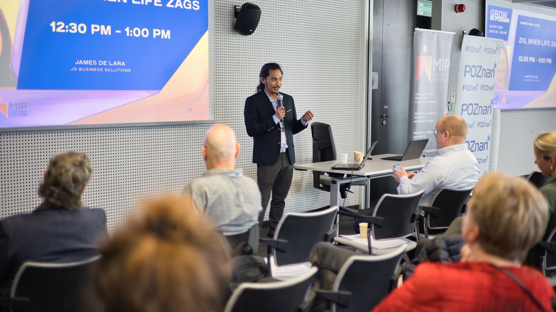 The speaker is addressing the audience during the conference. The audience and the displayed presentation are visible.