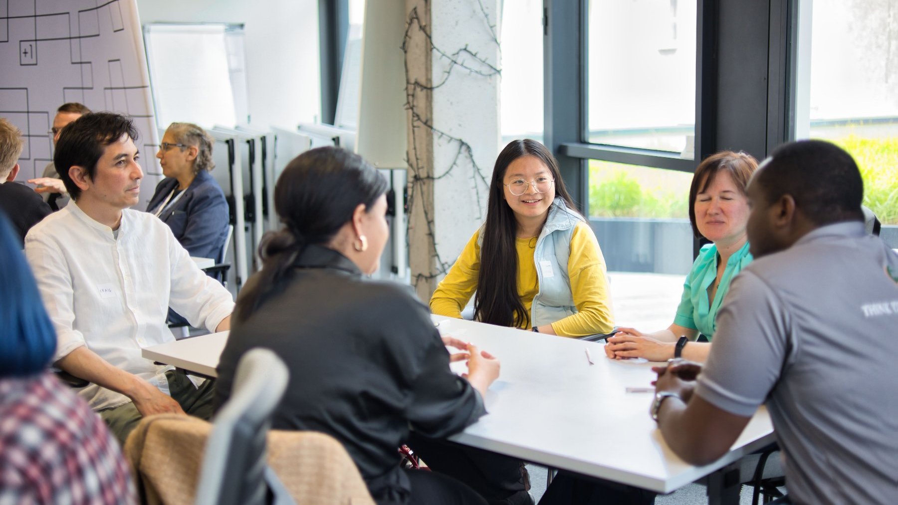 Five people are sitting at a table and discussing among themselves. Other teams can be seen around them. This is a networking workshop.
