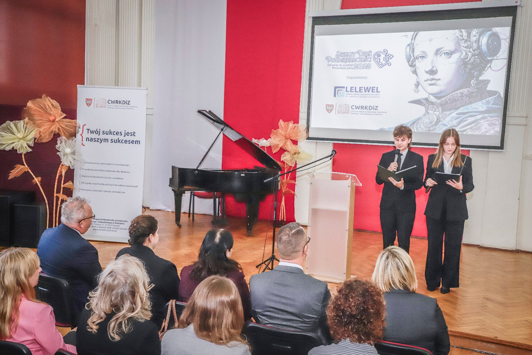 Aula szkolna, widoczni są goście audytorium oraz dwóch prowadzących uczniów.