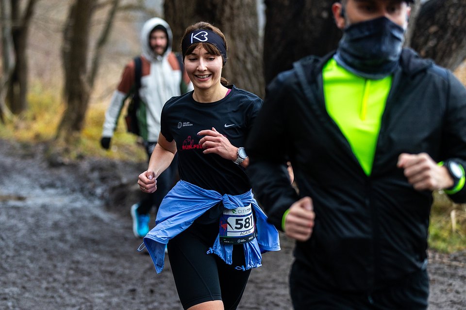 City Trail Poznań, fot. Tomasz Szwajkowski - na pierwszym planie biegnie uśmiechnięta w czarnych krótkich spodniach i krótkim rękawie. W tle widać idącego w kurtce mężczyznę, a z przodu biegnącego w czarnej kurtce mężczyznę
