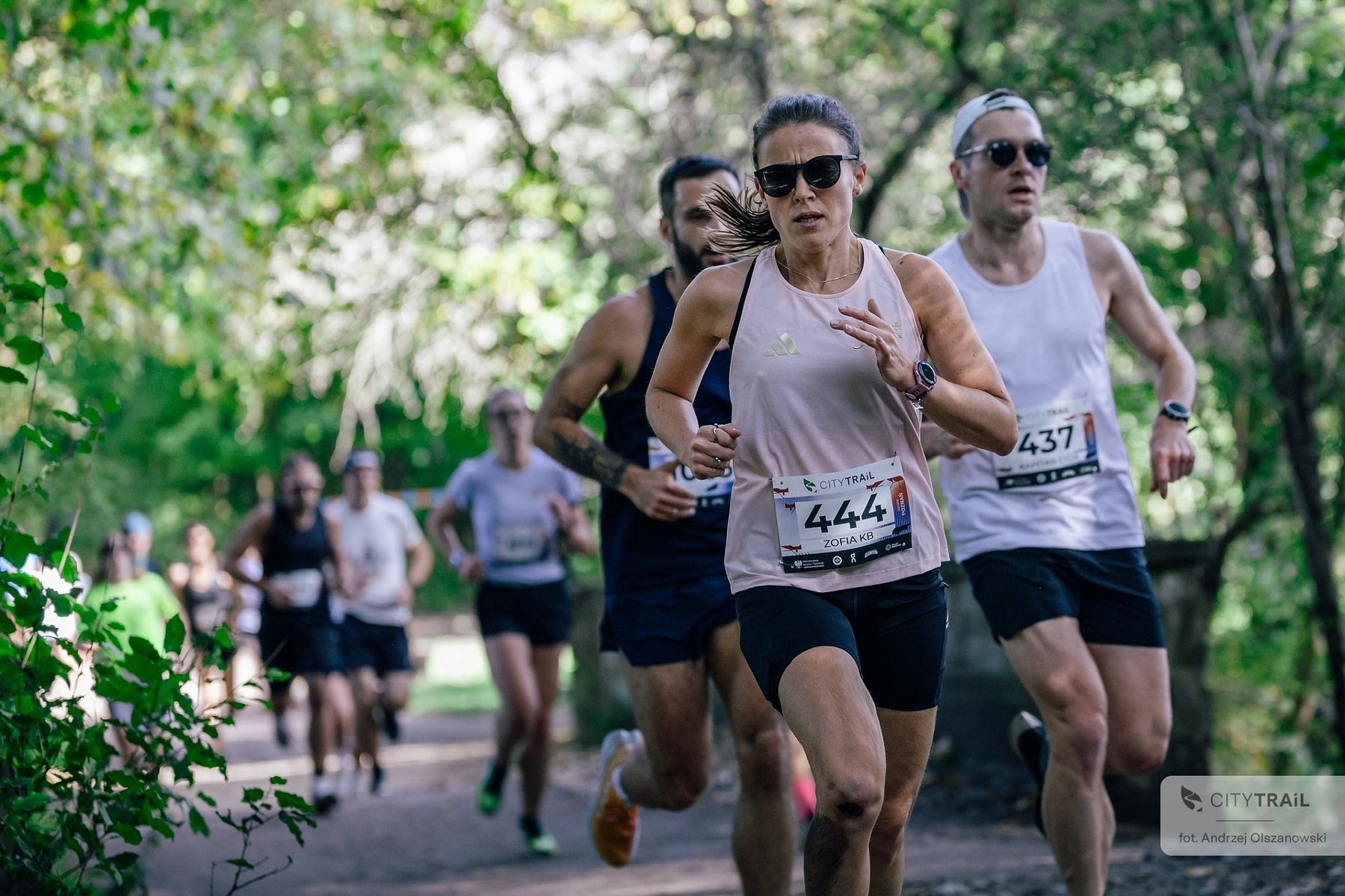 Cykl City Trail w Poznaniu, fot. Andrzej Olszanowski Cykl City Trail w Poznaniu, fot. Andrzej Olszanowski - na pierwszym planie trójka biegaczy w krótkich spodniach i koszulkach (1 kobieta oraz 2 mężczyzn) w tle kilkoro kolejnych biegaczy, a także drzewa z zielonymi liśćmi