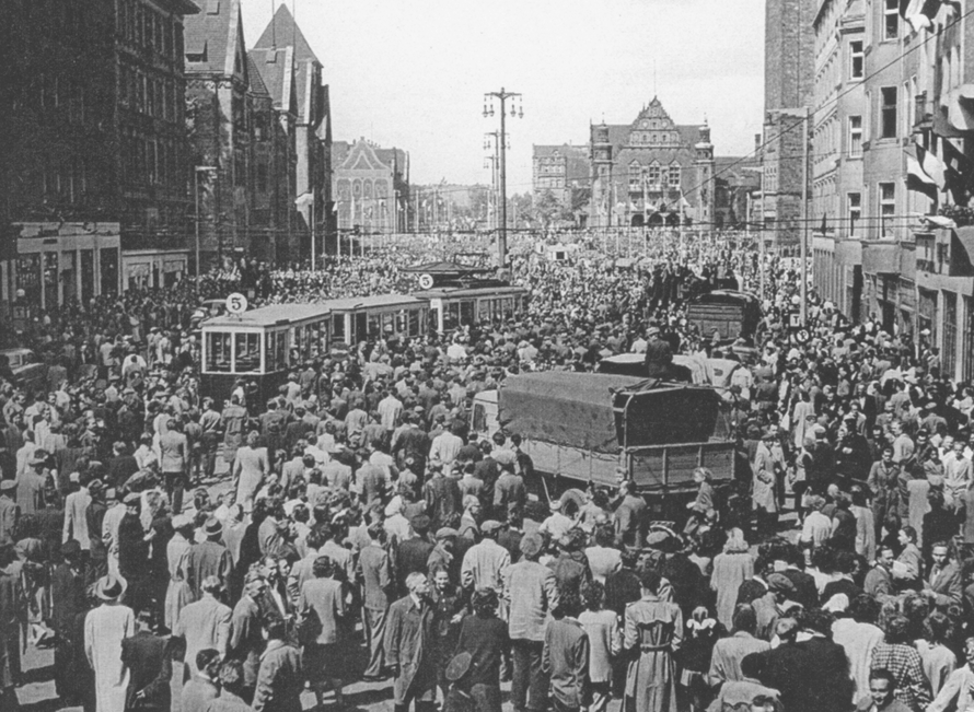 czarno białe archiwalne zdjęcie przestawiające tłum demontrantów w zabudowie śromiejskiej w tłumie samochodwy ciężarowe i tramwaje