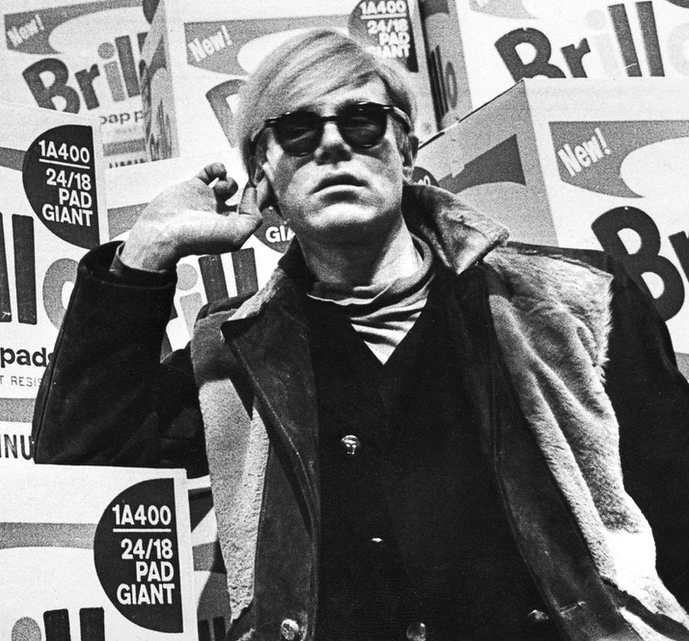 fot. Alfred Statler, "Andy Warhol in his studio at 1342 Lexington Avenue, New York City, April 1962", 1962, photographic negative / The Andy Warhol Museum, Pittsburgh; Museum Purchase © The Andy Warhol Museum, Pittsburgh, PA, a museum of Carnegie Institute. All rights reserved Czarno-białe zdjęcie Andy Warhola w okularach przeciwsłonecznych.