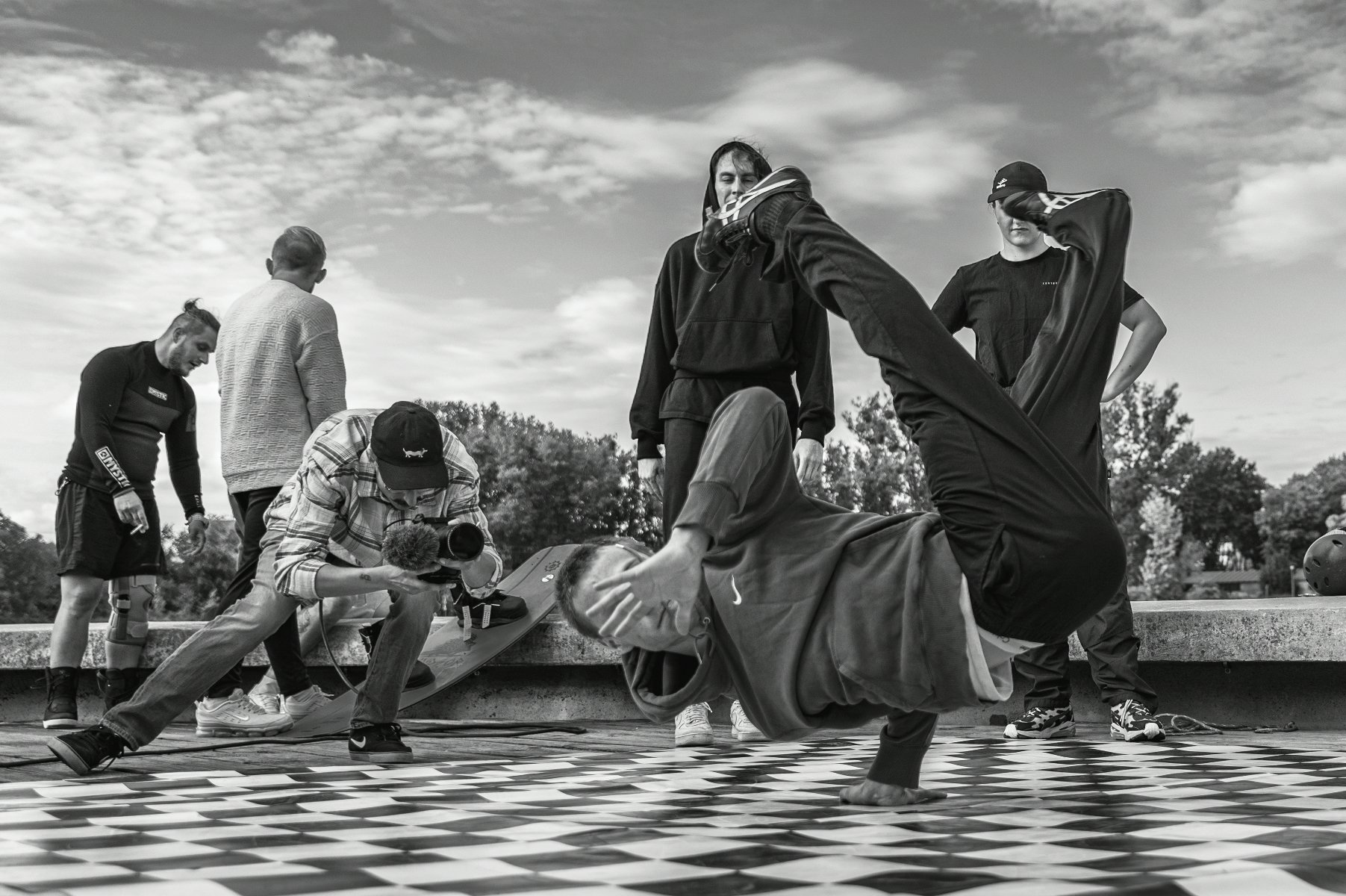 Czarno-białe zdjęcie przedstawiające grupę młodych mężczyzn na zewnątrz. Na pierwszym planie tancerz breakdance wykonuje akrobatyczny ruch, stojąc na dłoni na posadzce w czarno-białą kratę. Obok niego, inny mężczyzna klęczy, nagrywając jego występ kamerą. W tle stoi czterech innych obserwatorów.