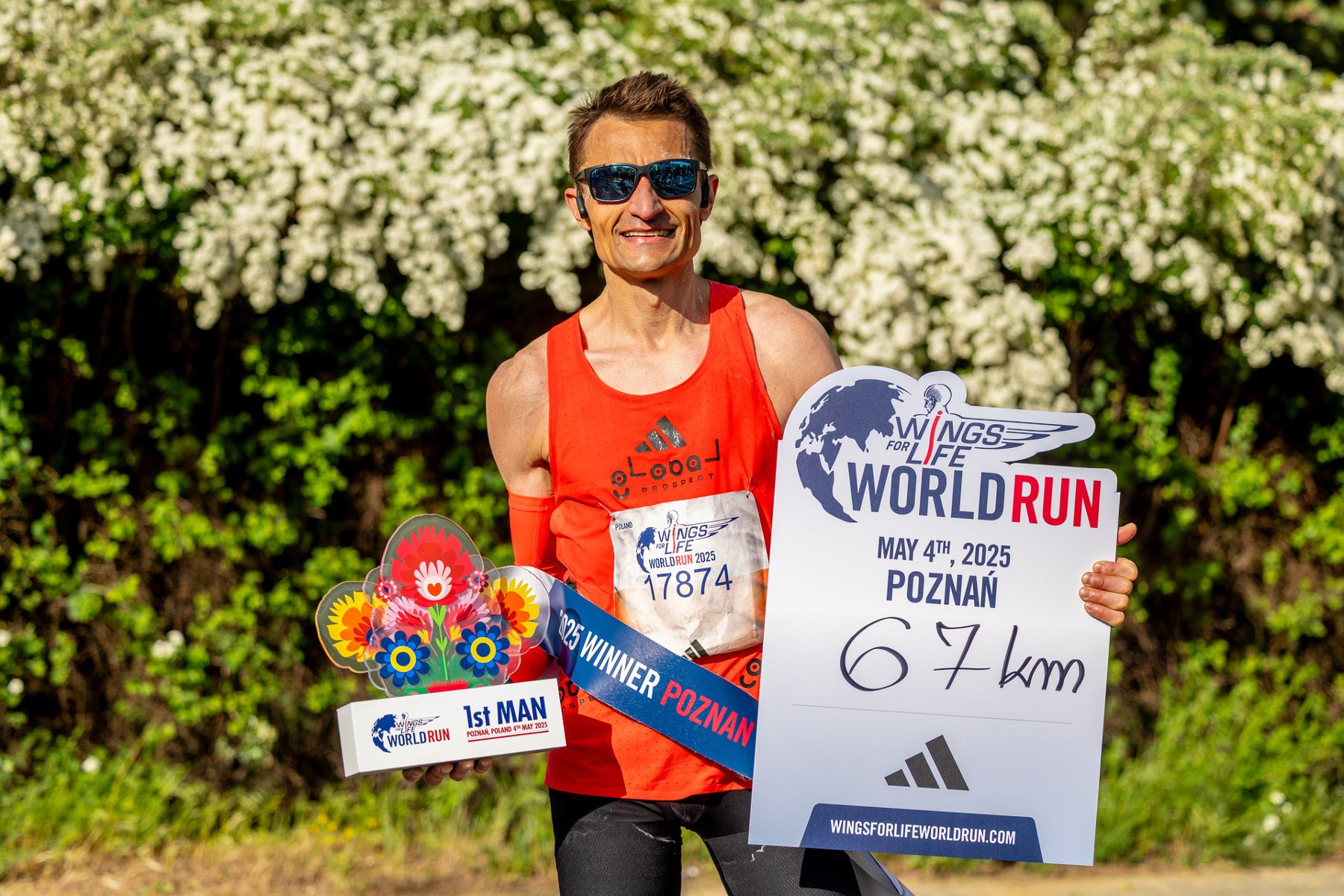 Dariusz Nożyński zwycięzcą biegu Wings for Life World Run w Poznaniu, fot. Damian Kramski Dariusz Nożyński zwycięzcą biegu Wings for Life World Run w Poznaniu, fot. Damian Kramski - mężczyzna stoi w czerownym stroju i okularach przeciwsłonecznych na tle zieleni i trzyma nagrodę za zwycięstwo w zawodach