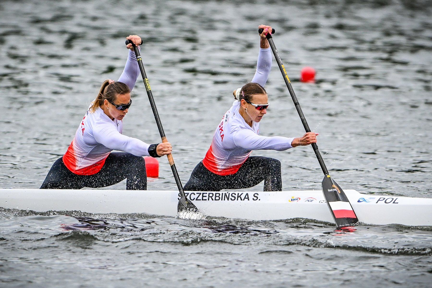 Dorota Borowska i Sylwia Szczerbińska na Torze Regatowym Malta, fot. Bartłomiej Zborowski Dorota Borowska i Sylwia Szczerbińska na Torze Regatowym Malta, fot. Bartłomiej Zborowski - 2 kobiety w biało-czerownych bluzach i czarnych spodniach płyną kajakiem po jeziorze