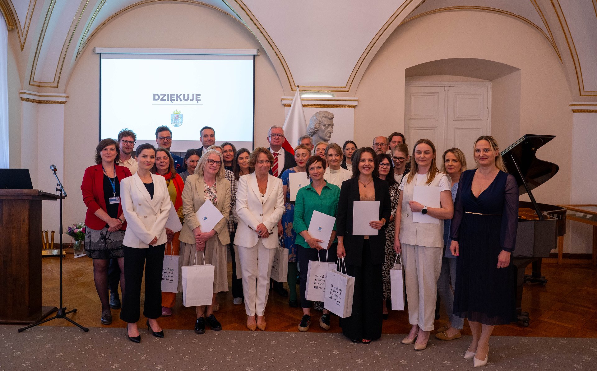 Biuro Miejskiego Konserwatora Zabytków w Poznaniu obchodzi jubileusz 70-lecia działalności Duża grupa ludzi, głównie kobiet w eleganckich strojach, pozuje do zdjęcia. W tle widoczny jest ekran z napisem "DZIĘKUJĘ", a także popiersie na postumencie i fragment fortepianu. Niektórzy z uczestników trzymają w rękach białe torby i certyfikaty.