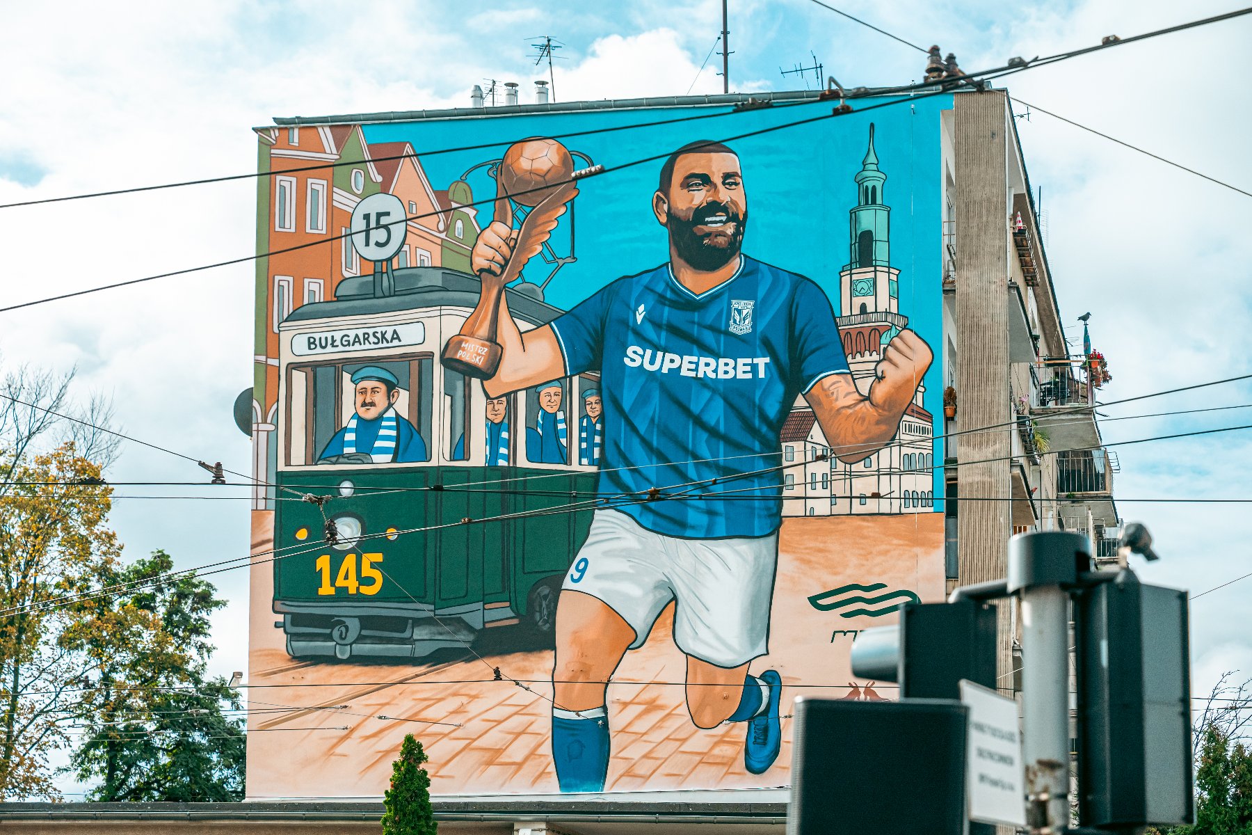 W stolicy Wielkopolski odsłonięto mural upamiętniający zdobycie przez Lecha Poznań mistrzostwa Polski w sezonie 2024/2025 Duży, kolorowy mural na bocznej ścianie budynku, namalowany w stylu komiksowym, przedstawiający sportowca i scenę z miasta. W centrum widać uśmiechniętego piłkarza w niebieskiej koszulce z napisem "SUPERBET" i numerem 9, który biegnie, trzymając w górze złotą statuetkę w kształcie piłki nożnej. W tle widoczny jest zielony tramwaj z numerem 145 i napisem "BULGARSKA", prowadzony przez motorniczego, oraz zabudowania miejskie (kamienice i Wieża Ratusza).