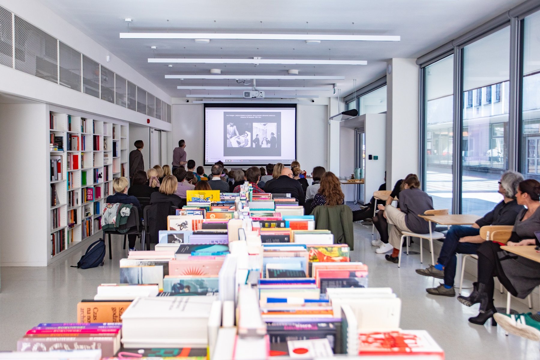 Fotografia przedstawia wnętrze Galerii Miejskiej Arsenał. Na środku stół zastawiony książakami, wokół ludzie siedzący przy stolikach na dalszym planie ekran rzutnika komputerowego.