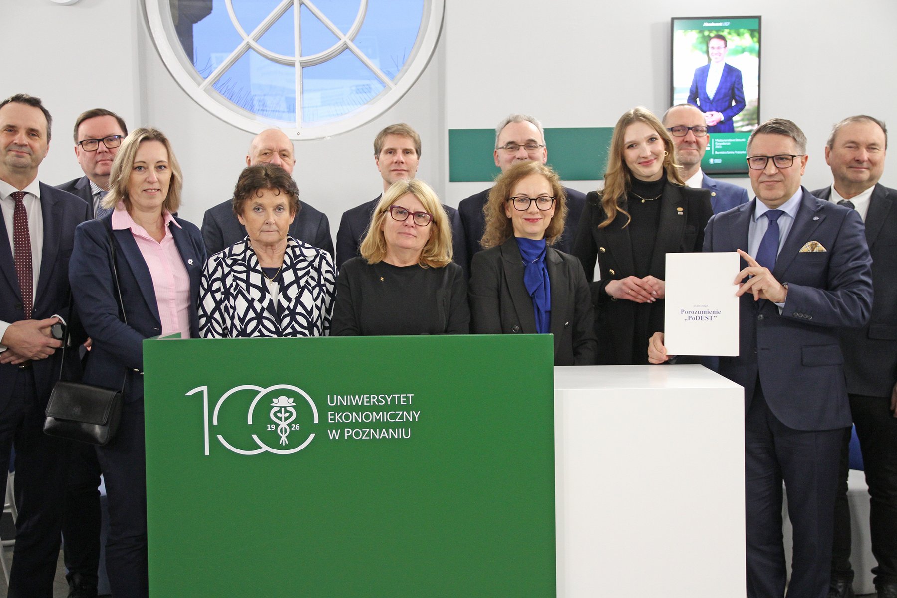 Grupa kilkunastu osób w strojach formalnych stoi za ladą z logo Uniwersytetu Ekonomicznego w Poznaniu; jedna z osób trzyma dokument porozumienia.
