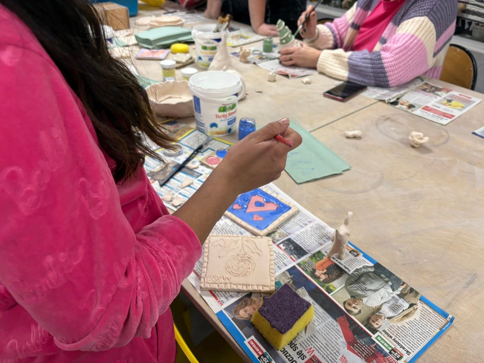 Grupa ludzi pracuje nad gliną przy stole, tworząc różne formy i rzeźby.