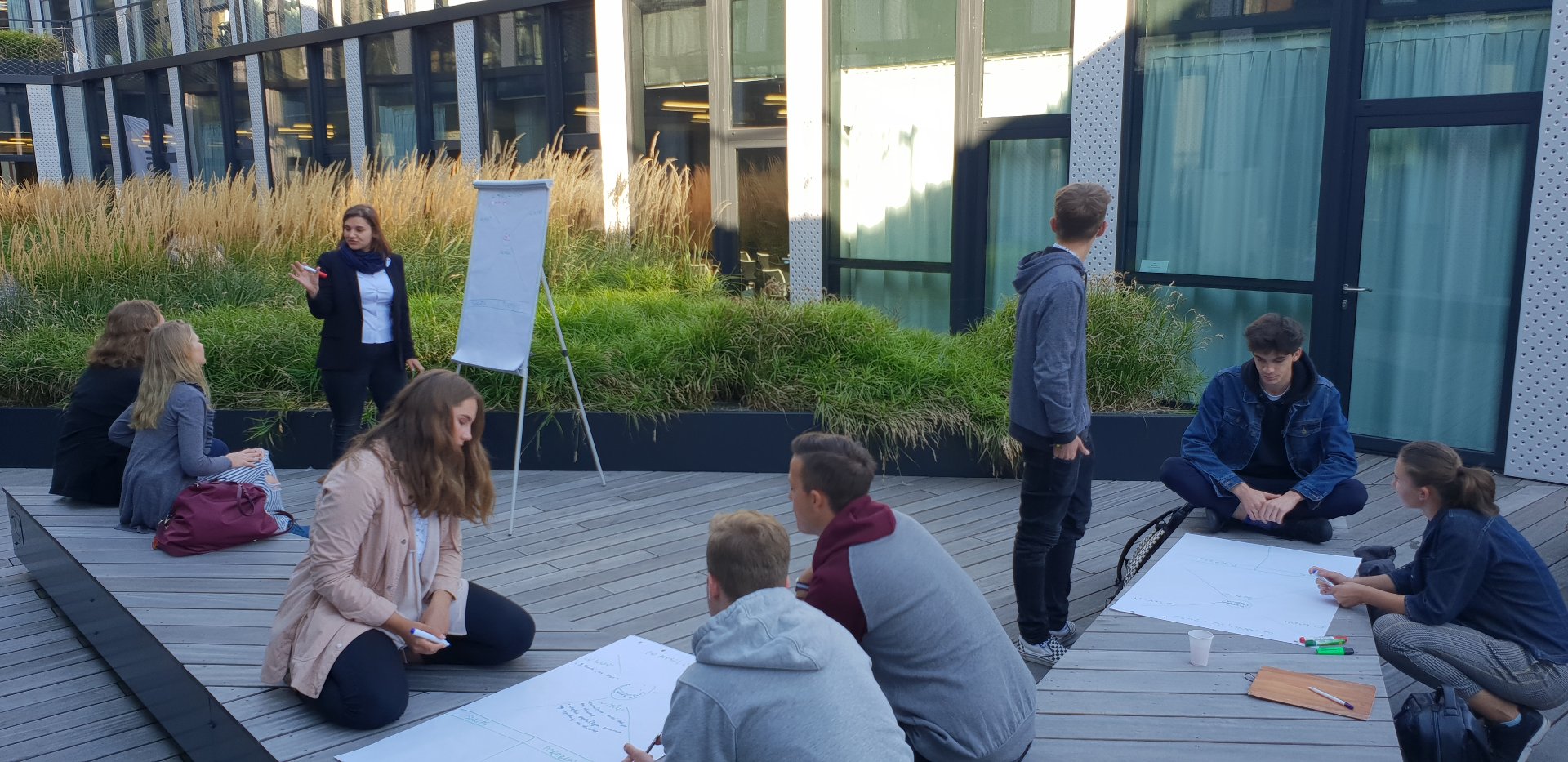 Grupa młodych osób pracuje w kilku zespołach na drewnianym tarasie przed nowoczesnym budynkiem; siedzą na ziemi, piszą na dużych kartkach, a jedna osoba stoi przy flipcharcie i tłumaczy