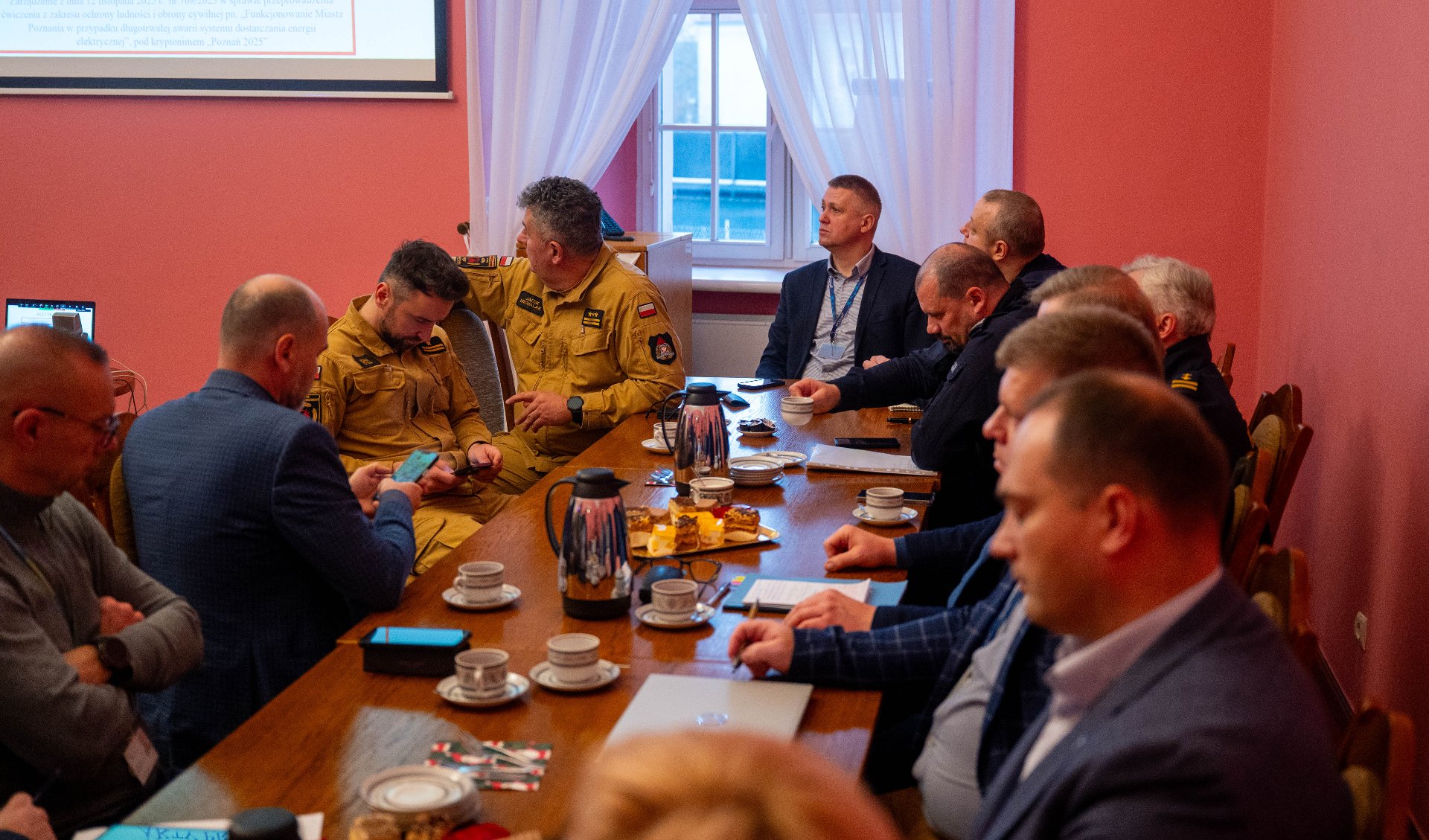 Grupa około dziesięciu mężczyzn w garniturach i mundurach siedzi przy dużym, drewnianym stole konferencyjnym w pomieszczeniu o czerwonych ścianach. Piją kawę i omawiają dokumenty.