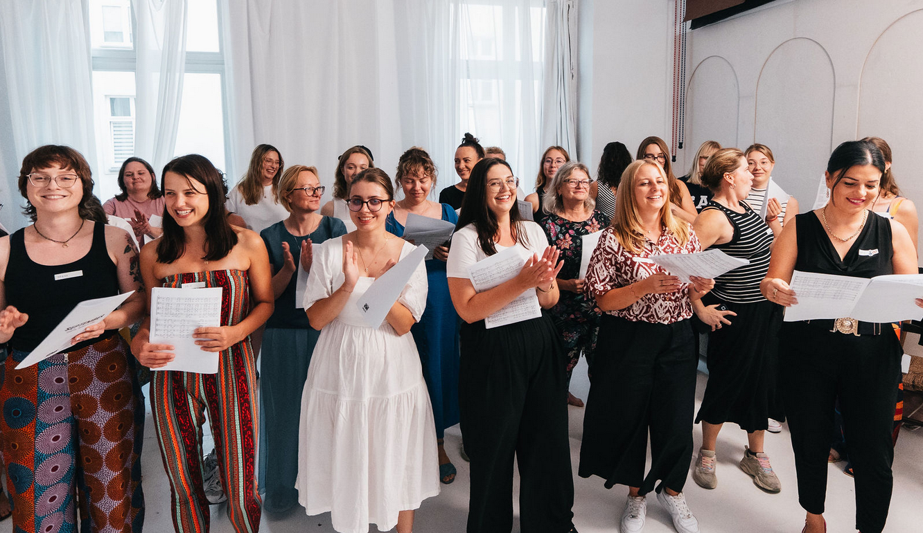 Grupa osób, która się uśmiecha i trzyma kartki z tekstem i nutami. Znajdują się oni w większej, białej przestrzeni. W tle widać okna i białe zasłony.