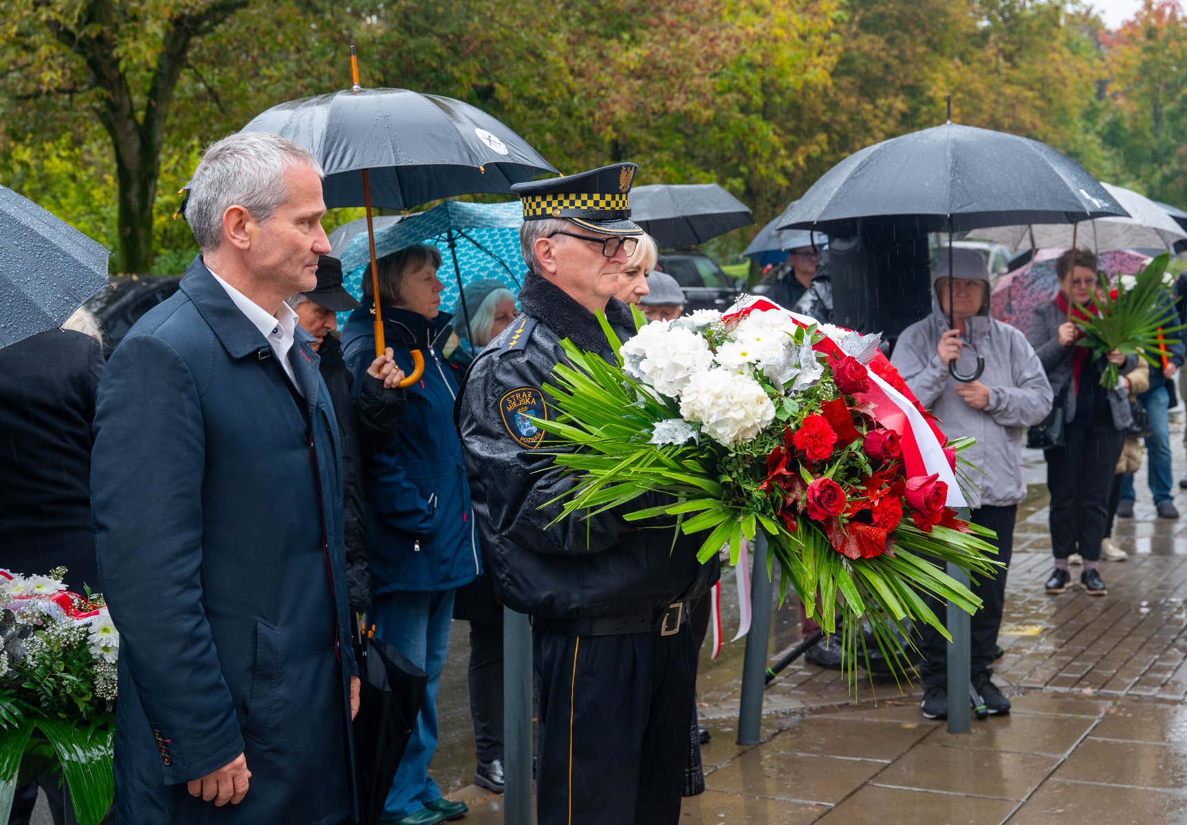 Grupa osób stoi na zewnątrz w deszczu pod czarnymi i niebieskimi parasolami, uczestnicząc w uroczystości pod pomnikiem. Na pierwszym planie widoczny jest mężczyzna w ciemnym płaszczu (po lewej) oraz drugi mężczyzna w czarnym mundurze i czapce ze złotym otokiem (pośrodku), który trzyma dużą, uroczystą wiązankę kwiatów złożoną z białych i czerwonych róż oraz białych hortensji z zielonym przybraniem, przewiązaną biało-czerwoną wstęgą.