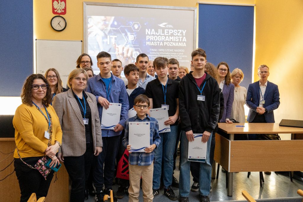 Poznaliśmy najlepszego programistę w Poznaniu Grupa uczniów w różnym wieku w sali lekcyjnej. Pozują z dyplomami i medalami, uśmiechnięci.